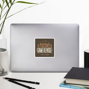 May include: A brown square sticker with a vintage illustration of a camper van in a forest setting. The text "GAMEHENEDGE" is in a retro font with the words "The art of escaping into nature" below it.