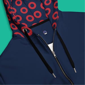 May include: A navy blue zip-up hoodie with a red circular pattern on the hood lining. The hoodie has a full zipper, black drawstrings, and a small embroidered design on the chest. The design is modern and casual.