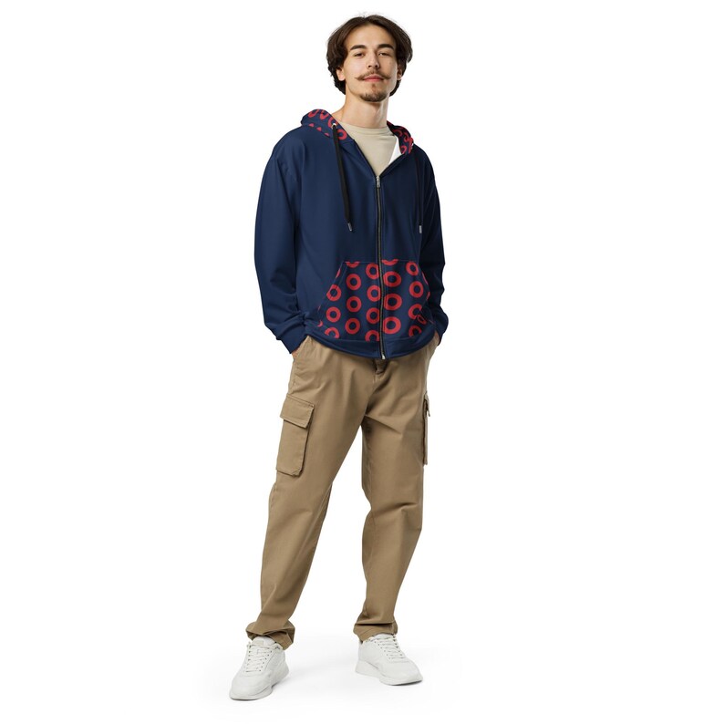 May include: A navy blue zip-up hoodie with a red donut pattern on the inside lining and pockets. The person is wearing khaki cargo trousers and white trainers. The hoodie has a full zipper and a drawstring hood.