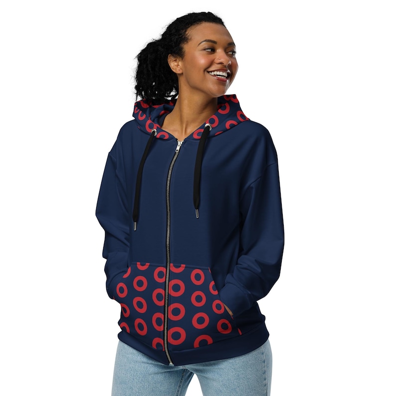 May include: Navy blue zip-up hoodie with a red circle pattern on the inside of the hood and pockets. The hoodie has a black zipper and drawstrings. The model is wearing blue jeans.