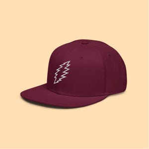 May include: A maroon baseball cap with a flat brim. The cap features a white, stylized lightning bolt design on the front. The hat is made of a soft material and has a structured fit.