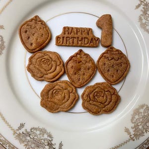 May include: A white plate with gold trim holds several brown cookies. The cookies are shaped like strawberries, roses, the number 1, and a banner that reads "HAPPY BIRTHDAY".