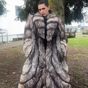 Puede incluir: Abrigo largo de piel de zorro plateado. El abrigo presenta un diseño lujoso y en capas con una mezcla de tonos plateados y grises oscuros. El modelo lleva botas negras. El abrigo es una pieza llamativa.