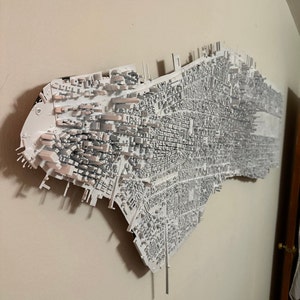 Könnte beinhalten: Ein weißes 3D-Modell der Skyline von New York, an einer Wand montiert. Das Modell ist detailliert und enthält viele der ikonischen Gebäude der Stadt.