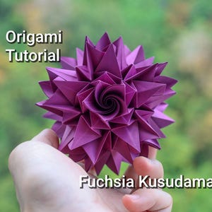 Puede incluir: Un kusudama de origami fucsia, una esfera de papel tradicional japonesa, se sostiene en una mano sobre un fondo verde borroso. El kusudama está hecho de muchas unidades de papel dobladas, creando una forma de flor.