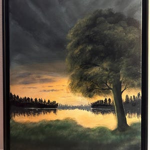 May include: Oil painting depicting a tranquil landscape with a large tree, a lake reflecting the sunset, and a dramatic, cloudy sky. The artwork is framed in a black frame and signed in the bottom right corner.