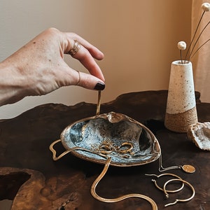 Puede incluir: Un joyero decorativo en forma de concha con detalles dorados, que contiene varios collares y pendientes dorados. Un pequeño jarrón blanco y marrón con flores secas está cerca. El plato está sobre una superficie de madera oscura.