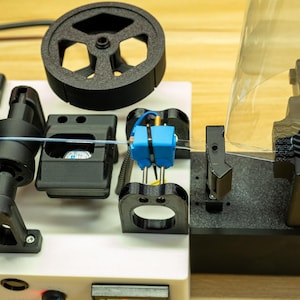 K&ouml;nnte beinhalten: Eine 3D-Drucker-Filament-Extrudiermaschine mit wei&szlig;er Basis, schwarzen mechanischen Komponenten, einem kreisf&ouml;rmigen Zahnrad und einem hellblauen Mittelblock. Ein d&uuml;nner, transparenter Filamentstrang verl&auml;uft horizontal durch die Mitte.