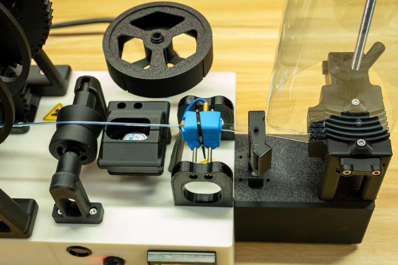 K&ouml;nnte beinhalten: Eine 3D-Drucker-Filament-Extrudiermaschine mit wei&szlig;er Basis, schwarzen mechanischen Komponenten, einem kreisf&ouml;rmigen Zahnrad und einem hellblauen Mittelblock. Ein d&uuml;nner, transparenter Filamentstrang verl&auml;uft horizontal durch die Mitte.
