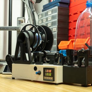 K&ouml;nnte beinhalten: Eine PET-Pultrusionsmaschine mit wei&szlig;em Geh&auml;use, digitaler Temperaturanzeige mit 230, blauem Drehknopf, schwarzem Filamentrollenhalter und einer transparenten Kunststoffflasche, die vertikal zur Materialzufuhr auf der rechten Seite montiert ist.