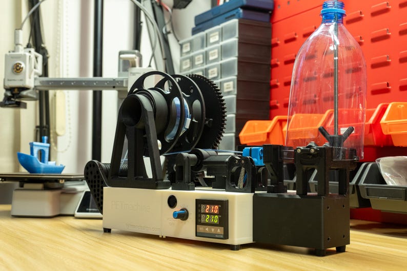 K&ouml;nnte beinhalten: Eine PET-Pultrusionsmaschine mit wei&szlig;em Geh&auml;use, digitaler Temperaturanzeige mit 230, blauem Drehknopf, schwarzem Filamentrollenhalter und einer transparenten Kunststoffflasche, die vertikal zur Materialzufuhr auf der rechten Seite montiert ist.