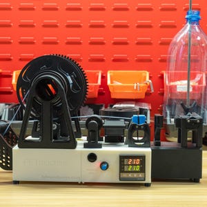 K&ouml;nnte beinhalten: Ein wei&szlig;-schwarzer PET-Maschinen-Kunststoff-Filament-Extruder mit einer digitalen Temperaturanzeige, die 210 Grad anzeigt, einem schwarzen Spulenhalter und einer klaren Plastikflasche auf einem vertikalen Stab zur Verarbeitung zu 3D-Druckmaterial.