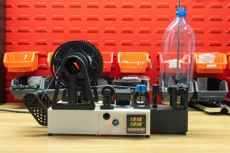 K&ouml;nnte beinhalten: Ein wei&szlig;-schwarzer PET-Maschinen-Kunststoff-Filament-Extruder mit einer digitalen Temperaturanzeige, die 210 Grad anzeigt, einem schwarzen Spulenhalter und einer klaren Plastikflasche auf einem vertikalen Stab zur Verarbeitung zu 3D-Druckmaterial.