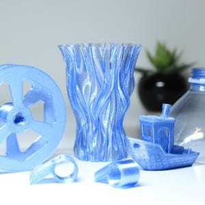 K&ouml;nnte beinhalten: Eine Sammlung hellblauer 3D-gedruckter Objekte, bestehend aus einem kreisf&ouml;rmigen Rad, einer strukturierten Vase, zwei kleinen ringf&ouml;rmigen Komponenten und einem Miniaturboot, neben dem oberen Teil einer transparenten Kunststoffflasche.