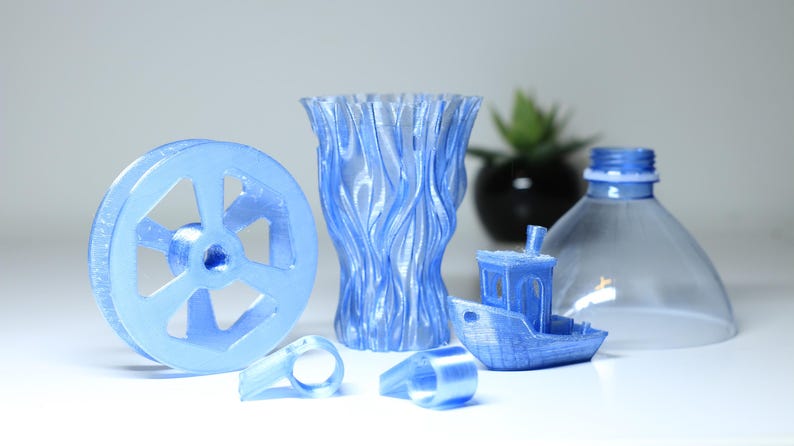 K&ouml;nnte beinhalten: Eine Sammlung hellblauer 3D-gedruckter Objekte, bestehend aus einem kreisf&ouml;rmigen Rad, einer strukturierten Vase, zwei kleinen ringf&ouml;rmigen Komponenten und einem Miniaturboot, neben dem oberen Teil einer transparenten Kunststoffflasche.