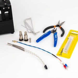 K&ouml;nnte beinhalten: Ein schwarzes elektronisches Ger&auml;t mit Digitalanzeige, zwei silberne Metallventile, ein kleiner Metallstift, d&uuml;nne Metalldr&auml;hte, ein Seitenschneider mit blauen Griffen, eine Zange mit schwarzen Griffen und eine gelbe Stanley-Klingenpackung.
