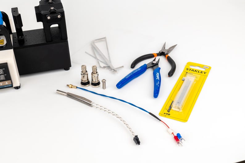 K&ouml;nnte beinhalten: Ein schwarzes elektronisches Ger&auml;t mit Digitalanzeige, zwei silberne Metallventile, ein kleiner Metallstift, d&uuml;nne Metalldr&auml;hte, ein Seitenschneider mit blauen Griffen, eine Zange mit schwarzen Griffen und eine gelbe Stanley-Klingenpackung.