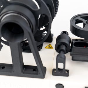 K&ouml;nnte beinhalten: Ein mechanisches Ger&auml;t mit wei&szlig;er Basis und schwarzen Komponenten, darunter ein gro&szlig;es Zahnrad, ein kreisf&ouml;rmiges Rad und ein Griff. Das Ger&auml;t enth&auml;lt zwei gelbe, dreieckige Warnschilder mit schwarzen Symbolen f&uuml;r Person und Hitze.