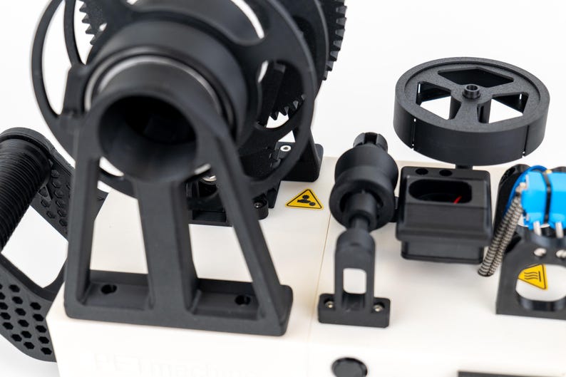K&ouml;nnte beinhalten: Ein mechanisches Ger&auml;t mit wei&szlig;er Basis und schwarzen Komponenten, darunter ein gro&szlig;es Zahnrad, ein kreisf&ouml;rmiges Rad und ein Griff. Das Ger&auml;t enth&auml;lt zwei gelbe, dreieckige Warnschilder mit schwarzen Symbolen f&uuml;r Person und Hitze.