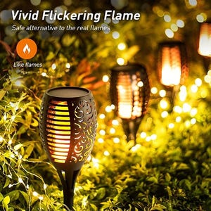 May include: A close-up of a solar powered flickering flame light. The light is black with a decorative design and emits a warm orange glow. The light is surrounded by green foliage and white string lights. The text "Vivid Flickering Flame" and "Safe alternative to the real flames" is displayed at the top of the image. The text "Like flames" is displayed below the light.