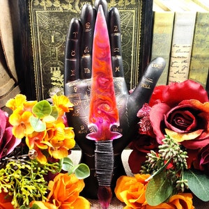 May include: A pink and orange resin dagger with a black cord wrap handle. The dagger is displayed on a black hand with astrological symbols.