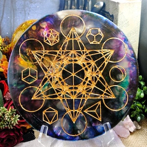 May include: A round, iridescent resin coaster with a gold metallic geometric design. The design features a star of David, a flower of life, and other sacred geometric shapes.