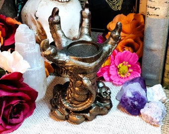 May include: A bronze-coloured candle holder shaped like a skeletal hand with a skull at the base. The hand is holding a small, round candle holder.