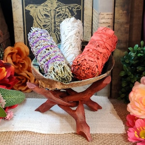 May include: Three bundles of smudge sticks, including white sage, lavender, and red palo santo, are displayed on a wooden stand. The stand is made of three branches that are connected in a tripod shape. The smudge sticks are arranged in a bowl-shaped container.