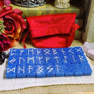 May include: A set of 24 blue rune stones with silver glitter and white runes. The stones are arranged in three rows of eight. The stones are in a red velvet pouch.