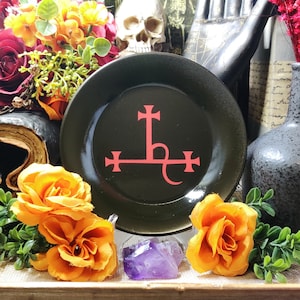 May include: A black ceramic plate with a red sigil of Baphomet. The plate is surrounded by orange flowers and a purple amethyst crystal.