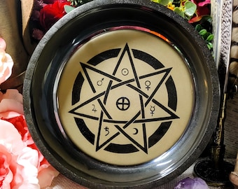 May include: A black slate dish with a gold rim and a black and white pentagram design with astrological symbols inside. The dish is on a white surface with flowers and crystals.