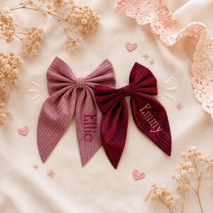 Può includere: Due fiocchi per capelli in tessuto a coste, uno rosa antico e uno bordeaux scuro. Il fiocco rosa antico presenta il nome ricamato Ellie, mentre il fiocco bordeaux scuro presenta il nome ricamato Emmy. Entrambi i fiocchi hanno code lunghe e appuntite.