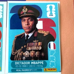 May include: A rectangular trading card featuring a portrait of a person in a dark military uniform with medals and a peaked cap. The card has a light blue background with the text DICTADOR MBAPPÉ, 20-12-1998, 1.82 m, 82 kg, and REAL MADRID CF.
