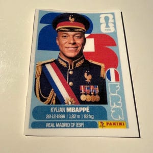 May include: A rectangular Panini trading card featuring Kylian Mbappé in a dark blue military-style uniform with gold epaulets, a sash, and medals. The background is light blue with the text 26, FRA, and the player's birth date, height, and weight.
