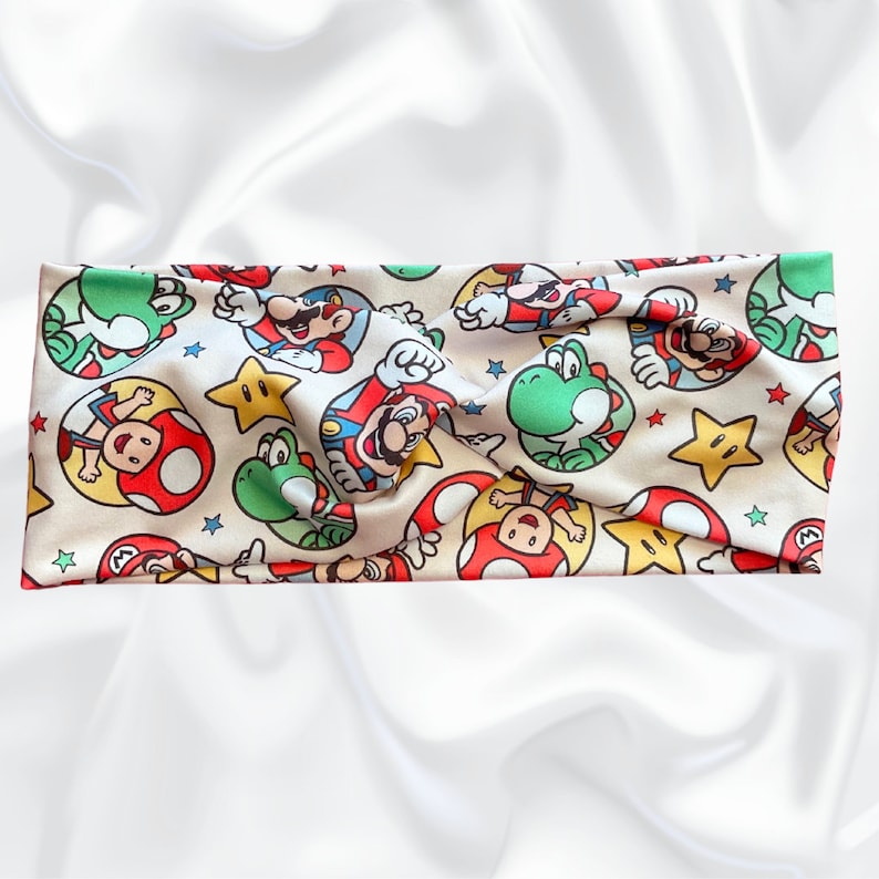May include: A beige headband with a repeating pattern of Mario, Luigi, Yoshi, and stars. The headband is made of a soft, stretchy fabric.