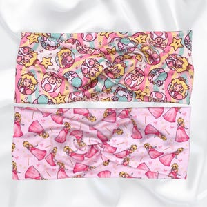 May include: Two pink and yellow patterned headbands. The top headband features a repeating pattern of Princess Peach, stars, and mushrooms. The bottom headband features a repeating pattern of Princess Peach in a pink dress.