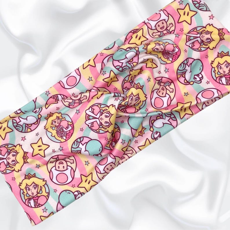 May include: A pink and green headband with a repeating pattern of stars, mushrooms, and a princess character. The headband is made of a soft, stretchy fabric.