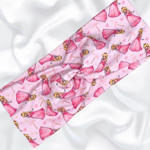 May include: Pink headband with a repeating pattern of a princess character in a pink dress and a crown. The princess is holding a wand and there are small pink bows and gold stars scattered throughout the pattern.