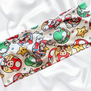 May include: A white headband with a repeating pattern of Mario, Luigi, Yoshi, and stars. The headband is made of a soft, stretchy fabric.