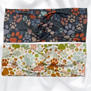 May include: Two twisted headbands with paw print patterns. One is dark gray with orange, peach, and teal paw prints. The other is white with orange, green, and peach paw prints. The headbands are made of a soft, flexible material.