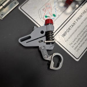 以下が含まれることがあります： 赤色のノブとスプリングが付いたグレーの機械部品。プリンターなどのデバイスの一部である可能性があります。部品には鍵穴型の開口部があり、印刷された指示の近くの暗い表面に配置されています。