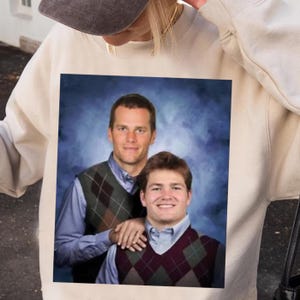 Puede incluir: Sudadera de color crema con una imagen impresa de dos hombres con chalecos de rombos y camisas con cuello sobre un fondo azul. La imagen parece ser un retrato. La sudadera es de color neutro.