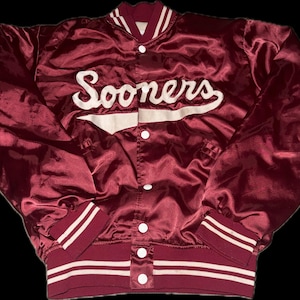 May include: Maroon satin jacket with the word "Sooners" in white script across the chest. The jacket has white stripes on the cuffs and hem, and a button-up front. A classic collegiate style.
