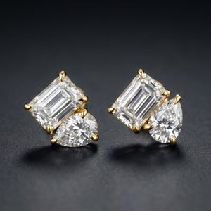 Puede incluir: Un par de pendientes de oro con diamantes. Cada pendiente presenta un diamante grande de talla rectangular y un diamante más pequeño en forma de pera. Los diamantes son claros y brillantes, sobre un fondo oscuro.