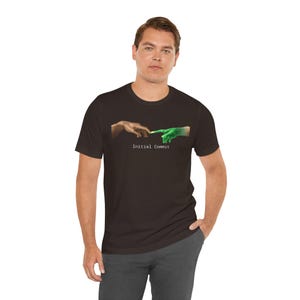 May include: Dark brown t-shirt featuring a graphic of two hands, one brown and one green, nearly touching. The design is a parody of the Creation of Adam, with the text "Initial Commit" below the hands.