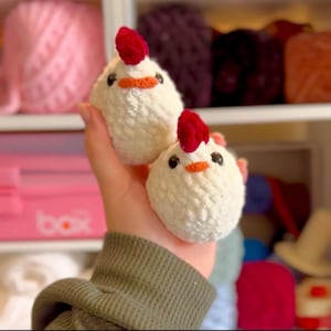 Op de afbeelding: Twee witte gehaakte kuiken speelgoed met rode en oranje accenten. De kuikens worden in een hand gehouden.