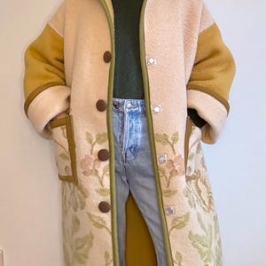 May include: A long beige coat with mustard yellow sleeves and collar. The coat has a floral pattern on the lower section and pockets, with brown buttons and green trim. Worn over a green top and blue jeans.