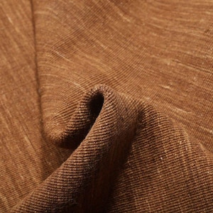 May include: Close-up of a textured brown fabric with a subtle, light-colored weave. The fabric is folded, creating shadows and highlighting the texture. The material appears to be a natural fiber, possibly linen or a similar textile.