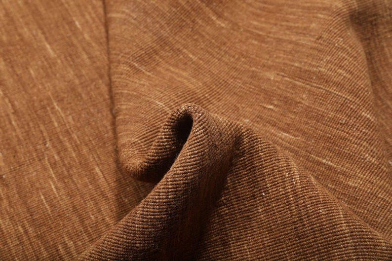 May include: Close-up of a textured brown fabric with a subtle, light-colored weave. The fabric is folded, creating shadows and highlighting the texture. The material appears to be a natural fiber, possibly linen or a similar textile.