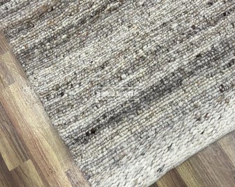 Op de afbeelding: Een getextureerd tapijt in neutrale tinten, met een geweven ontwerp. Het tapijt heeft een mix van grijze, beige en witte vezels, waardoor een subtiel, gevarieerd patroon ontstaat. De rand van het tapijt is een lichtere tint wit. Het tapijt ligt op een houten vloer.